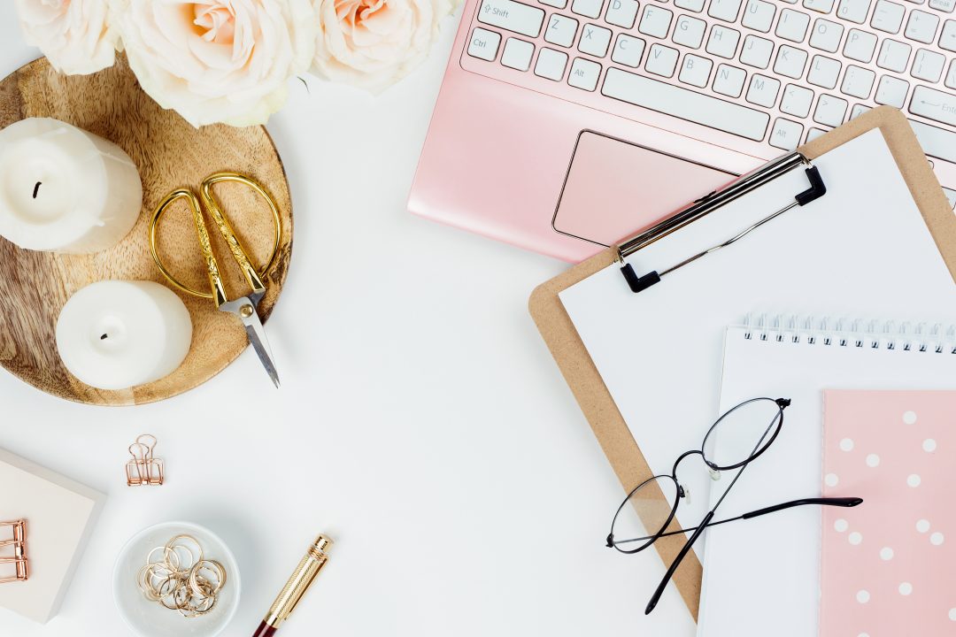 desk with blank open notepad, keyboard, stylish office / writing supplies and pink peonies on a white background, top view